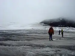 Drei Bergsteiger stehen auf einem dunklen Gletscher; rechts im Hintergrund erhebt sich ein schwarzer Berg.