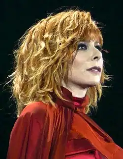 Mylène Farmer bei einem Liveauftritt 2019 mit lockigem, blondem Haar, die ein auffälliges rotes Outfit trägt. Sie schaut nach oben. Ausdrucksvoll und nachdenklich. Mylène Farmer trägt ein rotes Oberteil mit einem hohen Kragen, das durch einen darüber drapierten roten Stoff ergänzt wird. Der Hintergrund ist dunkel, wodurch die Person im Vordergrund besonders hervorgehoben wird. Die gesamte Szene wirkt stilisiert und dramatisch.
