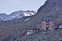 aus Stein gebaute Berghütte mit Nebengebäude im felsigen Gelände, im Hintergrund eine massive Bergwand, in höheren Lagen mit Schnee