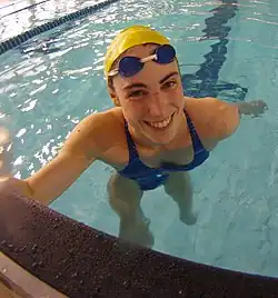 Anna Santamans befindet sich im Becken. Sie trägt eine gelbe Bademütze und einen blauen Badeanzug. Die Schwimmbrille hat sie auf die Stirn hochgeschoben.