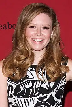 Natasha Lyonne mit langen, welligen, hellbraunen Haaren, die ein ärmelloses Kleid trägt. Das Kleid hat ein schwarz-weißes abstraktes Muster. Der Hintergrund ist ein kräftiges Rot und enthält teilweise sichtbaren Text mit der Aufschrift „Peabody.“ Das Foto wurde auf dem roten Teppich bei den Peabody Awards 2014 aufgenommen.