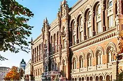 Eine seitliche Farbaufnahme des hellroten Backsteingebäudes mit seinen Rundfenstern und Säulen, verziert mit spitzen Türmen und Figuren. Die Architektur erinnert an barocke Kirchen.