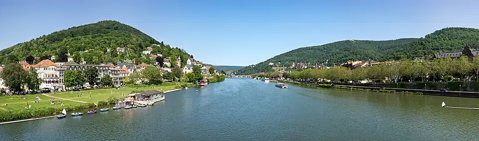 Blick von der Theodor-Heuss-Brücke ostwärts zum Ausfluss des Neckars aus dem Odenwald-Neckartal in die Oberrheinische Tiefebene mit der dortigen Alten Brücke in Heidelberg; links Heidelberg-Neuenheim mit der Neckarwiese und dem bewaldeten Heiligenberg-Vorgipfel Michelsberg, rechts hinter Bäumen die Heidelberger Altstadt und das bewaldete Königstuhlmassiv