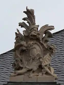 6. Trophäe – Baumstamm, Lanzenfahne, Trompete, Kartusche, Schild, Handspiegel, Rosen.