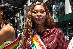 Porträtfoto von Jackson auf einer Pride Parade. Sie trägt ein dunkelrotes, faltiges Kleid, eine regenbogenfarbene Schärpe, eine goldene Halskette, rosa Lipgloss, aufgemalte Augenbrauen und goldroten Lidschatten. Ihre braunen Haare trägt sie auf der linken Seite nach hinten gekämmt, auf der rechten Seite bis zur Brust reichend und gewellt. Neben ihr steht eine schwarzhaarige Frau mit derselben Regenbogen-Schärpe, hinter ihr sind Zuschauer und Wohnhäuser mit Baugerüsten zu sehen.