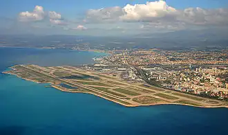 Farbfotografie in der Vogelperspektive von einem Flughafen mit Landebahnen, die links an drei Seiten vom Meer umgeben sind. Rechts ist eine große Stadt und im Hintergrund ist eine Berglandschaft.