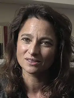 Nina Bouraoui (2016) mit langen, welligen Haaren und einem freundlichen Gesichtsausdruck. Sie befindet sich in einem Innenraum. Im Hintergrund ist ein Bücherregal sichtbar, in dem mehrere Bücher stehen. Die Beleuchtung im Raum wirkt angenehm und sanft, wodurch eine ruhige und gemütliche Atmosphäre entsteht.