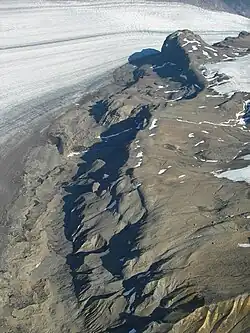 Luftbild eines spärlich mit Schnee bedeckten Berghangs mit einem Gletscher im Hintergrund