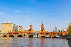 Frontale Farbfotografie von einer roten Backsteinbrücke mit einer gelben U-Bahn in der Mitte. Die Backsteinbrücke hat zwei große Türme und eine Unterführung für Autos und Fußgänger.