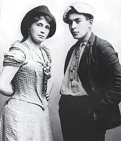 Schwarz-Weiß-Fotografie einer Frau und eines Mannes, die einander zugewandt im Viertelprofil in die Kamera sehen. Die Frau trägt ein gepunktetes Kleid, der Mann einen lässig geöffneten Anzug, beide haben Kappen auf dem Kopf.