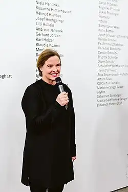 Brünette Frau mit Mikrofon vor einer Textwand mit Namen