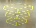 Die beiden oberen Pyramidenstümpfe scheinen über dem unteren Pyramidenstumpf, der scheinbar auf dem Fliesenboden steht, zu schweben. Tatsächlich handelt es sich aber nur um auf den Boden aufgeklebte gelbe Streifen.