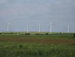 An area of flat green fields with five starkly white wind turbines standing out from the background of a blue sky.