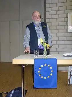 Tom Høyem in seiner Kluft als Wahlbeobachter bei einem Vortrag des Mittwochsforums Rheinstetten 2024. Er steht hinter einem Tisch, auf dem Blumen stehen und an dem eine kleine EU-Fahne hängt.