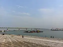 Foto einer sehr langen Brücke über einen breiten Fluss. Das sichtbare Ufer ist gepflastert. Auf dem Fluss sind einige Schiffe unterwegs.