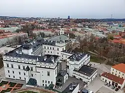 Farbfotografie in der Obersicht von einem weißen Schloss mit Seitenflügeln und schwarzem Dach. Links ist eine Kuppel und rechts ist ein gläserner Außenfahrstuhl. Im Hintergrund ist die Stadt mit Kirchen und einem Fernsehturm in der rechten oberen Ecke.
