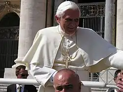 Frontale Farbfotografie von einem Papst mit einem goldenen Kreuz als Halskette, der sich an einem weißen Geländer festhält und seinen linken Arm lächelnd aus dem Foto streckt. Weiter unten und um ihn verteilt stehen Männer in schwarzen Anzügen.