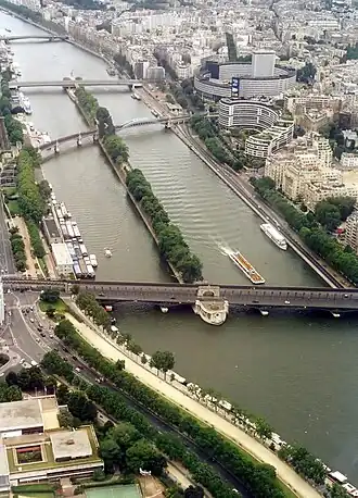 Farbfotografie in der Vogelperspektive von einem Fluss mit einer langen, schmalen Insel, auf der Bäume aufgereiht sind. Die Insel dient als Stütze für drei Brücken über den Fluss. Auf der vorderen Brücke fahren Autos und eine Eisenbahnbrücke führt über sie. Unten links und oben rechts sind viele Häuser und Uferalleen mit Schiffen.