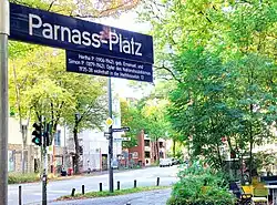 blaues Straßenschild mit Inschrift "Parnass-Platz" und Unterzeile mit einer kurzen Erklärung des Schicksals des Ehepaars Parnass