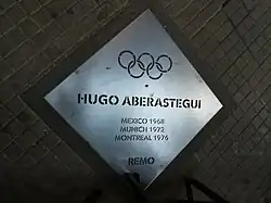 Auf der Tafel sind die Olympischen Ringe, der Name und die drei Olympiateilnahmen vermerkt. Darunter steht Remo, das spanische Wort für Rudern.