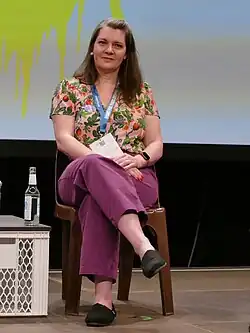 Foto einer Frau, die auf einem Plastikstuhl sitzt. Sie sitzt frontal vor dem Betrachter und hat ein Bein über das andere geschlagen. Sie trägt eine Mauvefarbene Hose und ein geblümtes Oberteil.