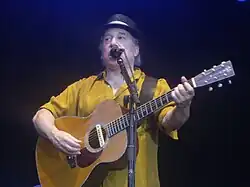 A man behind a microphone holding an acoustic guitar and wearing a yellow shirt and black hat.