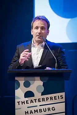 Paul Werner Glaser bei der Verleihung des Theaterpreises Rolf Mares 2023 in Hamburg. Er steht an einem Rednerpult und hält anscheinend eine Rede hält. Er trägt einen dunklen Anzug und ein weißes Hemd, was einen formellen Anlass suggeriert. Vor der Person befindet sich ein Mikrofon, das am Rednerpult befestigt ist. Auf dem Rednerpult ist ein Schild angebracht, auf dem der Text „THEATERPREIS HAMBURG“ deutlich zu lesen ist. Im Hintergrund ist ein blauer Bildschirm zu sehen, der mit weißen und blauen Kreisen gestaltet ist.