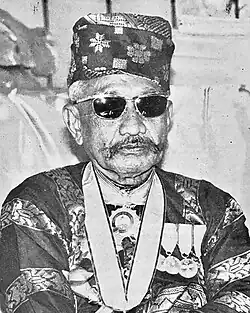 Pengiran Digadong Pengiran Anak Haji Khamis. Schwarz-Weiß-Porträt eines Asiaten mit zeremonieller Kopfbedeckung und großer Sonnenbrille.