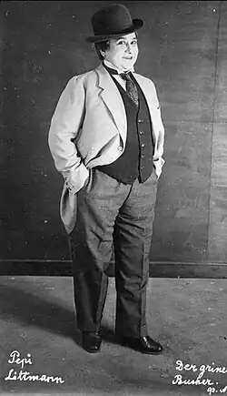 eine rundliche, weiblich-männliche Person im Hosenanzug mit Zylinder, Hände in den Hosentaschen, und verschmitztem Grinsen