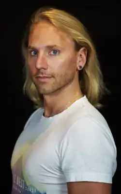 Robert Hankers (2023) mit schulterlangem, blondem Haar, der ein weißes T-Shirt trägt. Auf dem T-Shirt befindet sich ein farbenfrohes Grafikdesign mit dem Text „THE WEST COAST California“. Er trägt eine dünne Halskette und Ohrringe in beiden Ohren. Der Hintergrund des Bildes ist komplett schwarz, wodurch die helle Kleidung und das Haar der Person besonders hervorgehoben werden.