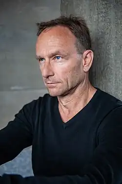 Dirk Schülke (2014) trägt ein schwarzes Oberteil. Er lehnt sich an eine Wand mit einer grauen, strukturierten Oberfläche im Hintergrund. Er hat kurzes, dunkles Haar und ist von der Brust aufwärts zu sehen. Abgesehen von der Person und der grauen Wand gibt es keine weiteren Elemente oder auffällige Objekte im Bild.