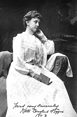 Die Groáufnahme von Wiggin zeigt, wie sie seitlich nach rechts gedreht vor dunklem Hintergrund auf einem Sofa sitzt. Sie trägt ein weißes Kleid und stützt ihren Kopf mit ihrem Arm auf der Sofalehne ab.