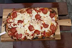 Ovales Gebäck ähnlich einer Pizza, auf dem man unter einer dicken Schicht weißem Mozzarella kleine Salamiräder erkennen kann. Die Pinsa liegt auf einem Holzbrett