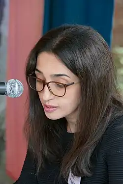 Porträtbild einer Frau mit langen dunklen Haaren und Brille, die in ein Mikrofon spricht. Blickrichtung ist nach links.