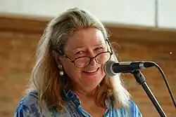 Porträt einer blonden Frau mit Brille, die den Kopf leicht schräg hält, in ein Mikrofon spricht und das Publikum ansieht.