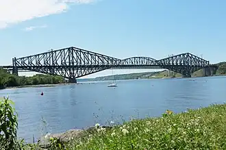 Das Bild zeigt die berühmte Pont de Québec, die größte Auslegerbrücke der Welt. Sie überspannt ein Gewässer, und im Vordergrund ist ein Segelboot zu sehen. Die Brücke besteht aus Stahl und hat eine komplexe Gitterstruktur, die ihre beeindruckende Größe und Stabilität betont. Sie verbindet zwei Landmassen, die von grüner Vegetation bedeckt sind. Der Himmel ist klar mit ein paar Wolken, und das Wasser ist ruhig. Zudem sind einige Bojen im Wasser sichtbar. Das Bild wurde von einer grasbewachsenen Stelle aus aufgenommen, die im Vordergrund auch einige Felsen und Pflanzen zeigt.