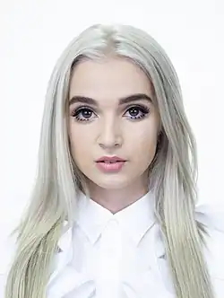 Moriah Rose alias Poppy mit langen, geraden, platinblonden Haaren und auffälligem Make-up, blickt direkt in die Kamera. Das Haar ist in der Mitte gescheitelt und fällt gleichmäßig auf beiden Seiten des Kopfes bis über die Schultern. Sie trägt ein weißes Hemd mit einem hohen Kragen und Rüschen-Details an den Schultern. Der Hintergrund ist komplett weiß, wodurch die hellen Haare und das Hemd besonders hervorgehoben werden. Das Bild vermittelt eine klare und minimalistische Ästhetik durch den Kontrast zwischen den Haaren und der Kleidung.