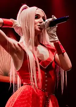 Sängerin Poppy bei einem Auftritt auf einer Bühne. Sie trägt ein auffälliges rotes Kleid, das mit silbernen Knöpfen und Verzierungen geschmückt ist. Das Kleid hat einen tiefen Ausschnitt und einen weit ausgestellten Rock, der einen dramatischen Eindruck vermittelt. Ergänzt wird das Outfit durch weiße Handschuhe und rote Armbänder, die einen eleganten Touch hinzufügen. Sie hat lange, blonde Haare, die offen über die Schultern fallen. Sie hält ein Mikrofon in der rechten Hand und steht in einem Bereich mit roter Bühnenbeleuchtung, die eine intensive, dramatische Atmosphäre erzeugt. Der Hintergrund ist dunkel gehalten, wodurch die Sängerin durch die Beleuchtung noch stärker hervorgehoben wird.