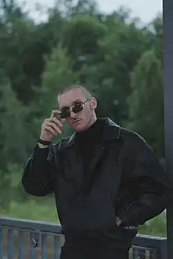 Almin Bejic mit kurzen Haaren trägt schwarze Sonnenbrille und schwarzen Mantel, steht draußen vor grünem Hintergrund