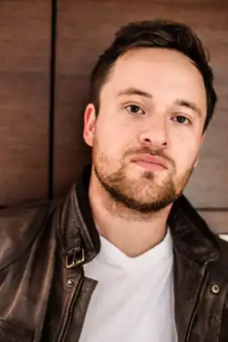 Portrait des deutschen Schauspielers Lorris Andre Blazejewski im Jahre 2018. In brauner Lederjacke, darunter ein weißes Shirt, an eine braune Wand gelehnt und ernst in die Kamera schauend. Foto geschossen von Fotografin Sandra Nauheimer.