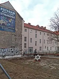 Foto des Grundstücks mit der Baulücke nach dem Abriss des Vorgängergebäudes, ein weißer Davidstern (Durchmesser etwa 1 Meter) wurde mitten auf die Freifläche gestellt, an der Brandmauer des Nachbarhauses hängt ein riesiges Poster: „Potsdam baut eine Synagoge. Helfen Sie mit! Spenden Sie! Synagogen-Förderverein Potsdam e.V.“