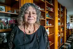 Eine Farbfotografie zeigt Rita Laura Segato stehend im Porträt. Sie hat halblanges, lockiges, grause Haar und schaut direkt in die Kamera. Sie trägt ein graues Oberteil und verschiedene Ketten.