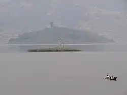 Das Foto im Querformat zeigt eine wenige Quadratmeter große Insel in einem See. Diese ist von Gras und einem einzelnen kleinen, recht kahlen Baum bewachsen. Vor der Insel rechts paddeln in einiger Entfernung zwei Männer in einem Einboot davon. Im trüben Hintergrund befindet sich ein größerer Hügel, vermutlich auf einer weiteren Insel, und dahinter das steile Seeufer mit einzelnen, nicht genauer zu erkennenden Häusern, Baumstreifen und Feldern.