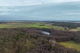 Ein Luftbild einer Naturlandschaft aus Wald, Wiesen und einem See, mit einer relativ kleinen, vierseitigen Natursteinpyramide mit hellen Eingangstor, die auf einer kleinen Waldlichtung inmitten eines Mischwald steht. Die Pyramide ist nicht höher als die Bäume.