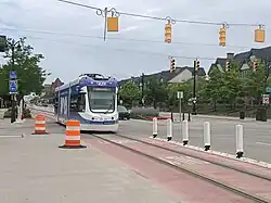 Straßenszene unter bewölktem Himmel. Straßenbahn links im Bild, verschiedene Straßenmöblierung wie Verkehrsleittonnen und Flexposts, darüber Lichtsignalanlagen an Spanndrähten. Im Hintergrund niedrige Häuser hinter Straßenbegleitbäumen.