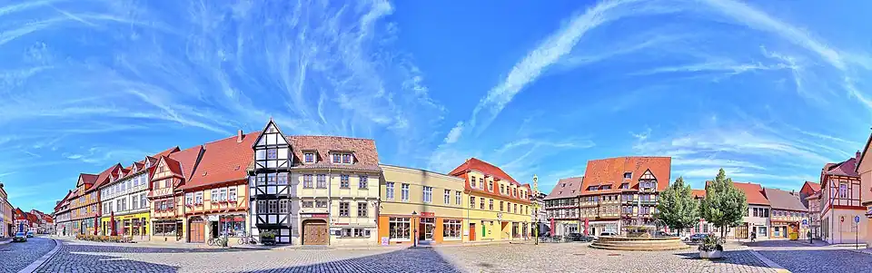 Panorama des Neustädter Markts in Quedlinburg mit Fachwerkhäusern, Kopfsteinpflaster und einem Brunnen unter blauem Himmel