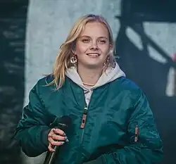 Paula Hartmann lächelt, während ihres Auftritts beim Rocken am Brocken 2022. Sie trägt eine grüne Bomberjacke über einem grauen Kapuzenpullover. In ihrer rechten Hand hält sie ein Mikrofon, während sie scheinbar inmitten eines Songs oder einer Ansprache ist. Ihre blonden Haare fallen locker um ihr Gesicht, und sie trägt auffällige Ohrringe. Der Hintergrund ist verschwommen, aber eine Bühne und Bühnenbeleuchtung sind erkennbar, was die Konzertatmosphäre unterstreicht.