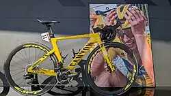 Das gelbe Canyon Aeroad mit der Startnummer 11 von Katarzyna Niewiadoma, mit der sie den Gesamtsieg der Tour de France 2024 holte. Am Lenker hängt noch ihr Helm. Dahinter ist überlebensgroß das sehr emotionale Foto von dem Moment, als sie unmittelbar nach dem Rennen total erschöpft erfährt, dass sie mit nur 4 Sekunden Vorsprung den Gesamtsieg errungen hat.