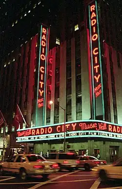 An einer Verkehrsstraße steht ein graugrünes Hochhaus, im Erdgeschoss befindet sich ein braunes Schild mit hellblauer Umrandung. Auf dem Schild steht der rote Schriftzug Radio City Music Hall, darunter in weißen Buchstaben das Theaterprogramm. Über dem Schild im Erdgeschoss sind zwei hohe, längliche Schilder mit derselben Aufschrift angebracht. Am Gebäude hängen einige Landesflaggen, während auf dem Gehsteig Fußgänger und eine Straßenlaterne stehen. Auf der Straße fahren zudem einige Autos.