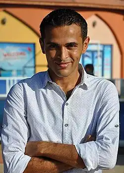 Rami Aman. Porträt eines jungen dunkelhäutigen Mannes im Hemd mit aufgekrempelten Ärmeln und verschränkten Armen.